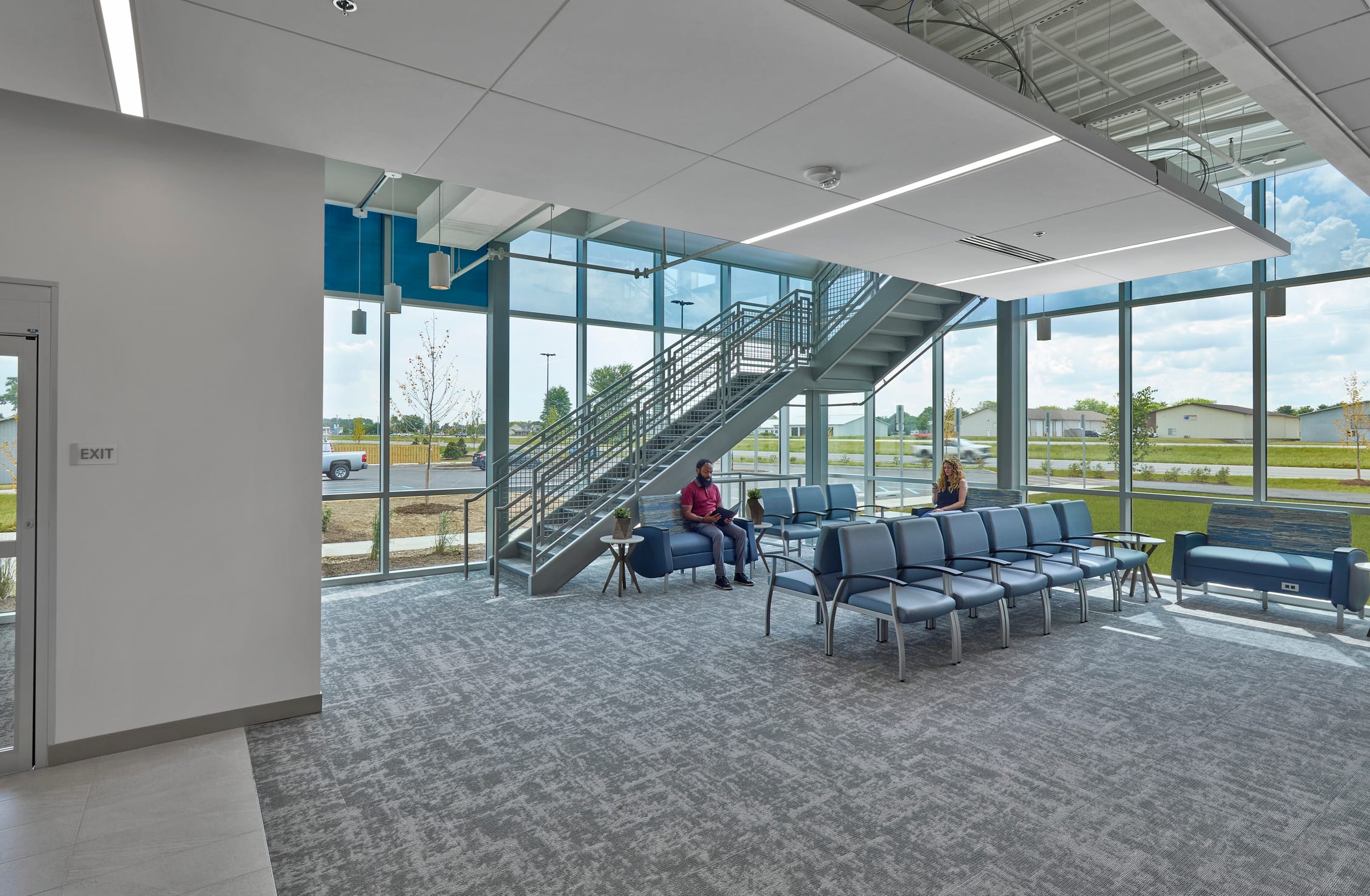 This image shows a waiting area in the Southern Indiana Orthopedics facility, featuring a dramatic two-story space with expansive floor-to-ceiling windows that offer views of the surrounding landscape. A prominent metal staircase with industrial-style railings connects to an upper level. Two people sit conversing in blue chairs near the windows, while additional rows of blue seating are arranged throughout the space. The area uses consistent gray carpeting that matches the previous interior image, maintaining the facility's cohesive design aesthetic. Natural light floods the space, creating an airy, open environment. The architectural design emphasizes transparency and connection to the outdoors while providing a comfortable waiting environment for patients and visitors. An exit door is visible on the left wall, and the exposed ceiling structure above adds to the contemporary architectural statement.