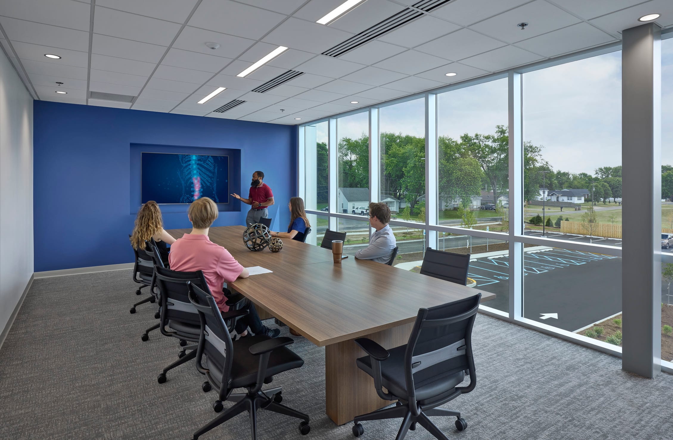 The interior of Southern Indiana Orthopedics in Columbus, IN, reflects a modern, professional, and patient-centric design approach. This meeting or training room showcases a clean, bright atmosphere with floor-to-ceiling windows that bathe the space in natural light while offering views of the surrounding community. The color palette is crisp and calming, with a bold blue feature wall that anchors the room and adds visual interest. The built-in digital screen, set into a recessed niche, hints at the space's technological integration and focus on education and communication—fitting for a facility dedicated to orthopedic care. Furnishings are contemporary and comfortable, with ergonomic chairs and a long wooden conference table that promotes collaboration. Overall, the design promotes transparency, wellness, and an environment of focused professionalism.