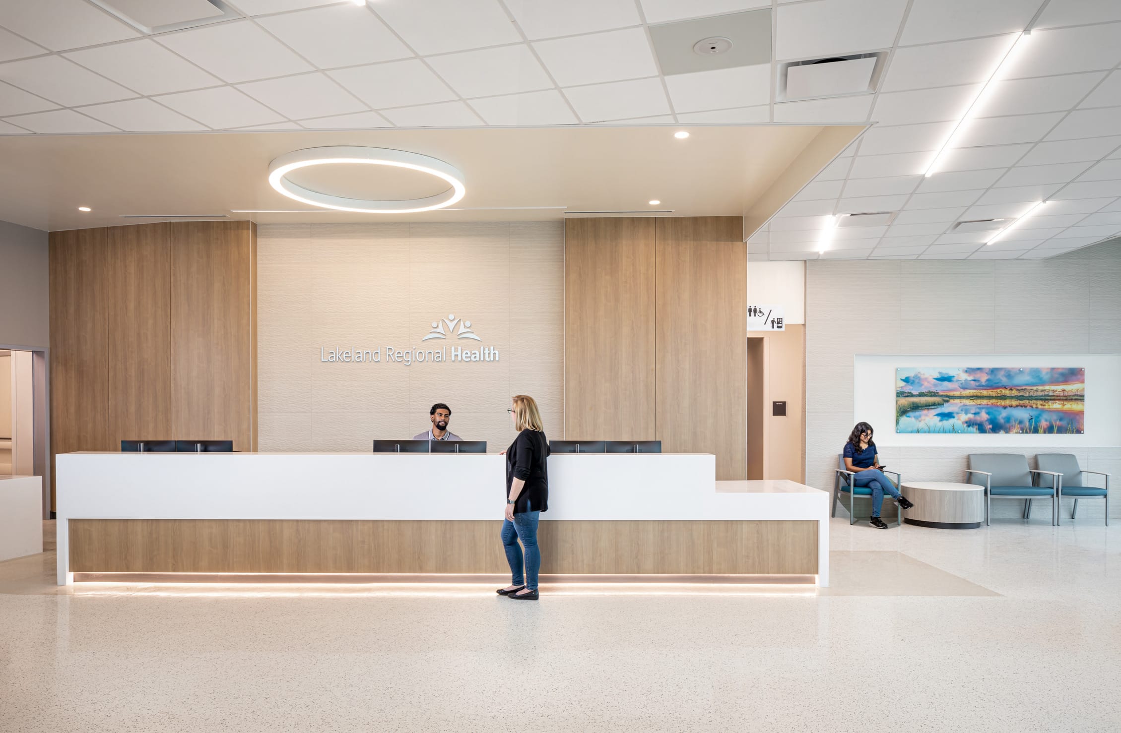 Modern reception area of Lakeland Regional Health featuring a sleek white and wood-toned welcome desk with subtle under-lighting. A staff member assists a woman in black top and jeans at the counter. Behind the desk, the facility's logo is displayed on a textured wall beneath a circular ceiling light fixture. To the right, a small waiting area includes blue-gray chairs, a round coffee table, and a landscape artwork with water reflections. The space combines contemporary design with warm materials, creating a welcoming healthcare environment with polished floors and recessed lighting.