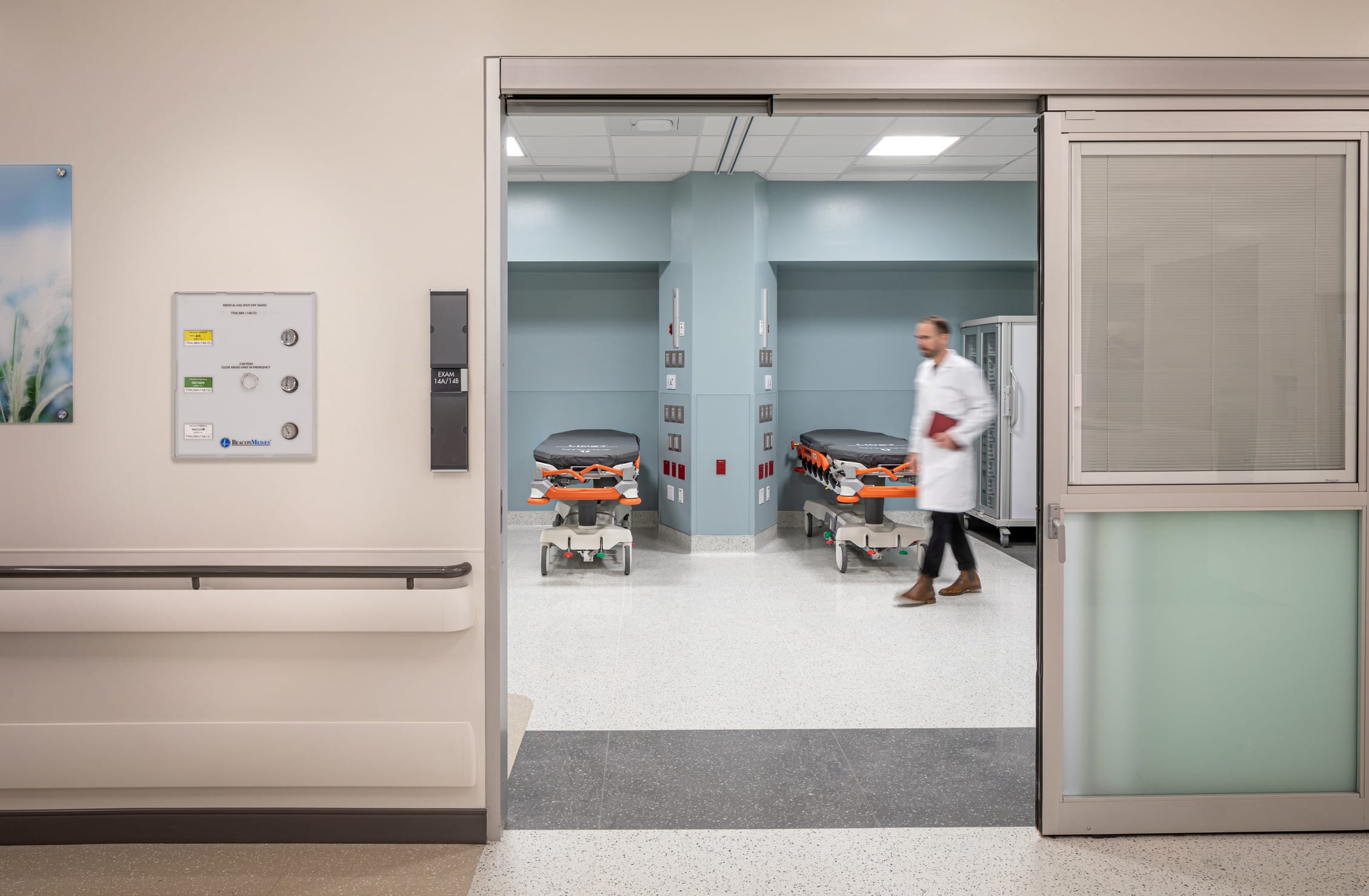 Interior view of an emergency department treatment area showing an open doorway leading to a room with two transport gurneys with orange safety straps. A healthcare professional in a white coat carrying a clipboard walks through the space. The pale blue walls feature medical equipment mounts and control panels. To the left of the doorway is a wall-mounted control panel and a handrail. The floor is speckled white with a contrasting darker threshold strip. The clinical environment is clean, modern, and designed for efficient emergency care.
