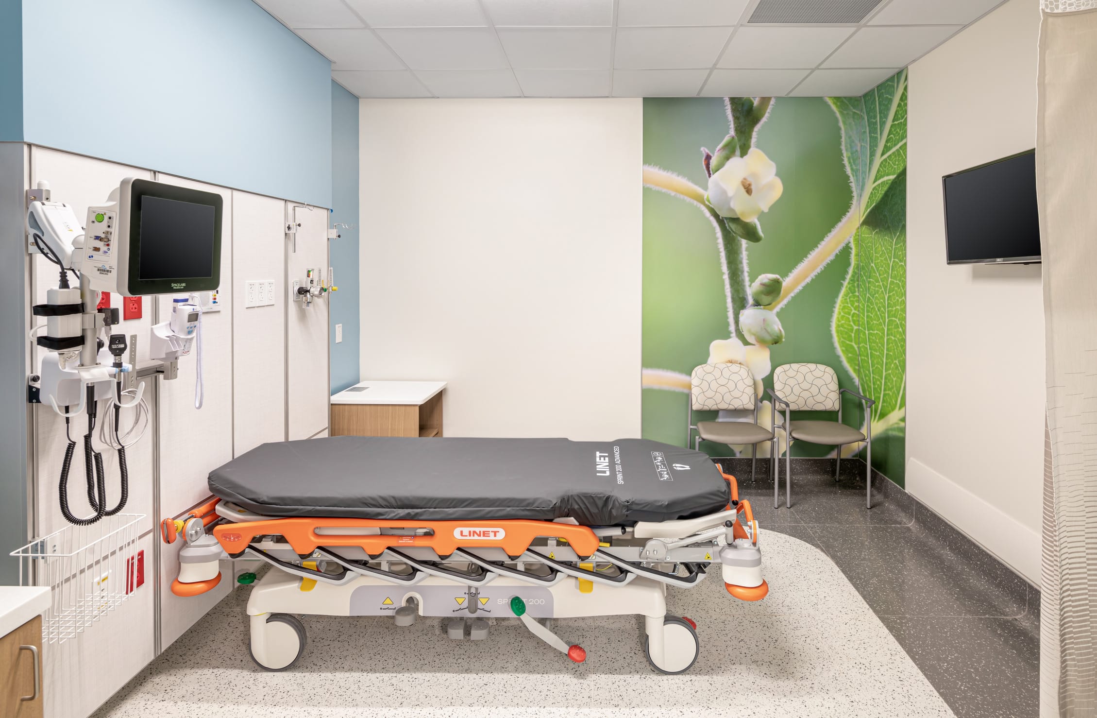 Emergency treatment room featuring a LINET adjustable medical gurney with gray mattress and orange safety rails. Wall-mounted medical equipment including vital signs monitor and examination tools are positioned to the left. The room is brightened by a large nature-themed wall mural showing a close-up of plant stems and buds, creating a calming atmosphere. Two patterned visitor chairs sit against the mural wall, and a wall-mounted television is visible in the upper right corner. The space combines clinical functionality with biophilic design elements to reduce patient stress in the emergency care environment.