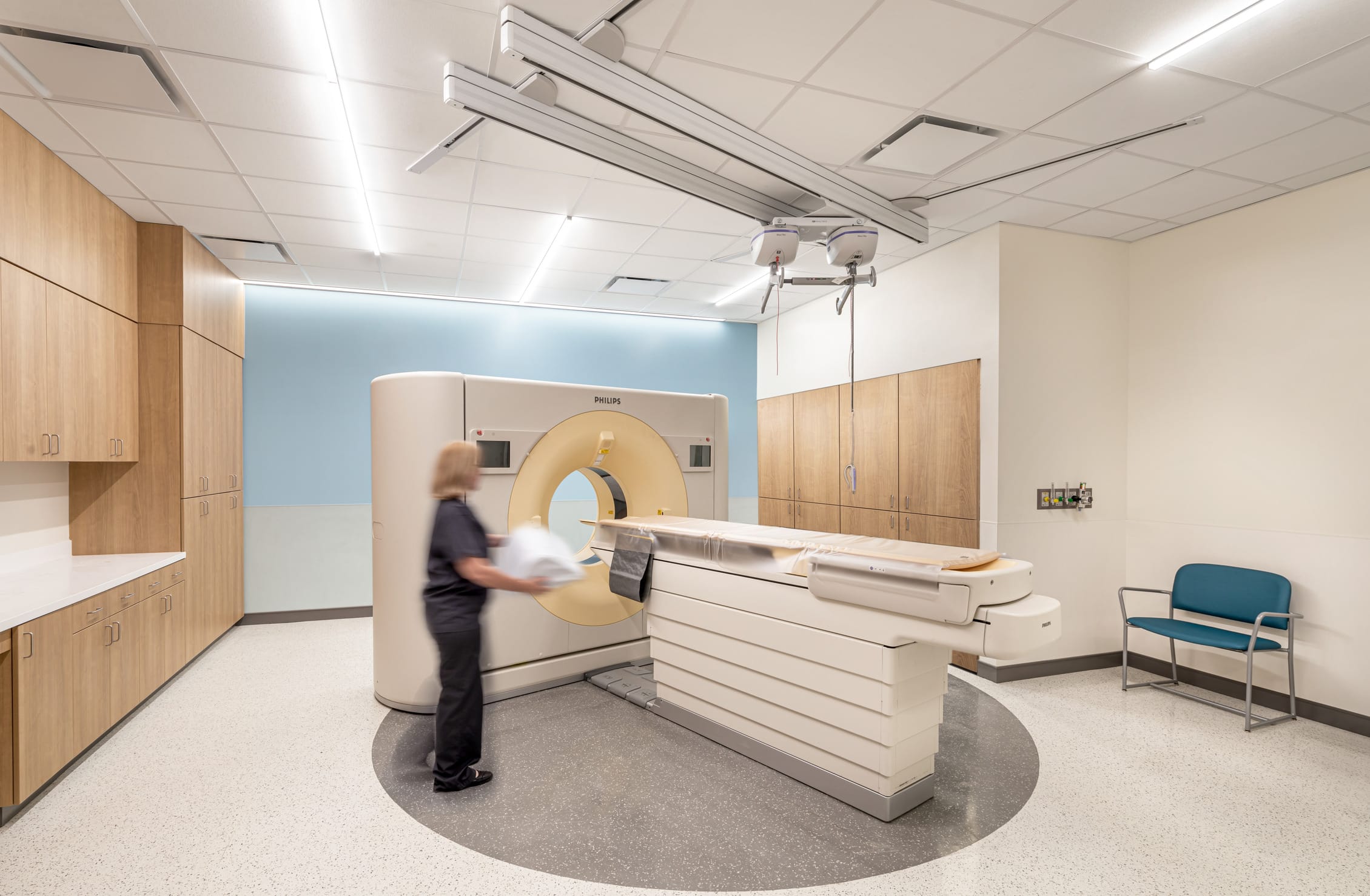Medical imaging room featuring a Philips CT scanner with its distinctive circular gantry and patient table positioned in the center of the room. A technician in dark clothing stands beside the machine, preparing for a procedure. The room has a calming blue accent wall, warm wood cabinetry along two walls, and built-in storage. Ceiling-mounted equipment rails and lighting provide functionality above the scanner. A teal chair sits in the corner for accompanying persons. The floor features a circular inlay marking the scanner's position. The space is designed for efficient workflow while maintaining a less clinical atmosphere.